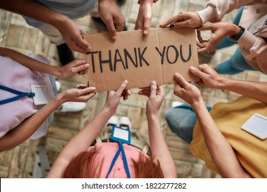 Top view of diverse young volunteers holding card with Thank you lettering while standing in charitable organization office, Selective focus on hands and inscription, Horizontal shot - Powered by Shutterstock