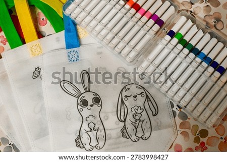 Top view of a DIY creative set for children featuring white eco-friendly textile bags with cute bunny outlines for coloring. Next to the bags is a large open plastic case with a variety of colorful felt-tip pens and markers. Educational hobby and art therapy concept for kids.