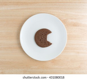 Top View Of A Dutch Cocoa Cookie That Has Been Bitten On A White Plate Atop A Wood Table Top.