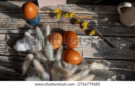 Top view Easter arrangement with brown eggs, soft pastel bunny tail stems, a white bunny ornament, and blooming pussy willow branch of goat willow (salix caprea) on distressed wooden boards. Delicate spring styling with natural seasonal texture.