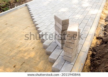 Top view of the edge of surface with laid gray paving slabs. Surface with sandy mixture is visible. Two stack of paving slabs is ready for laying.