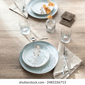 Top view of an elegantly set dining table, light blue ceramic plates, simple glassware and woven coasters, set on a rustic wooden table textile napkins, floral decor, Art Deco-inspired dining ambiance - Powered by Shutterstock