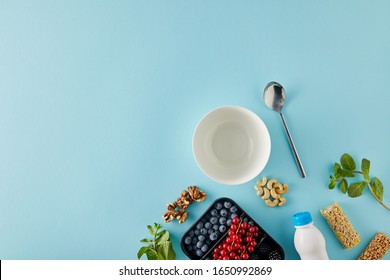 Top View Of Empty Bowl With Spoon, Container With Berries, Bottle Of Yogurt, Nuts, Cereal Bars, Mint On Blue Background