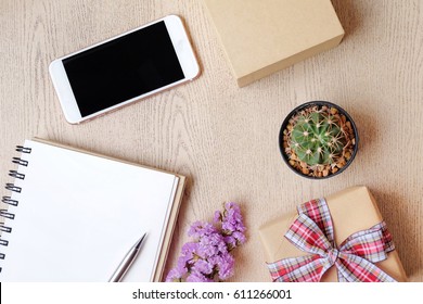 Top view of empty notebook, cell phone, cactus pot, flower bouquet  and gift box on white wood table.