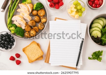 Top view of empty notebook with pen and healthy food including grilled chicken roasted potatoes broccoli asparagus salad berries and cheese on white marble table