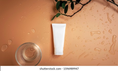 Top view empty organic facial skincare white tube product next to shining glass cup with branch and leaves on droplets orange brown background.