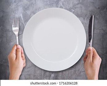 Top view of empty white ceramic plate with hand with knife and fork isolated on dark table gray cement wallpaper background.