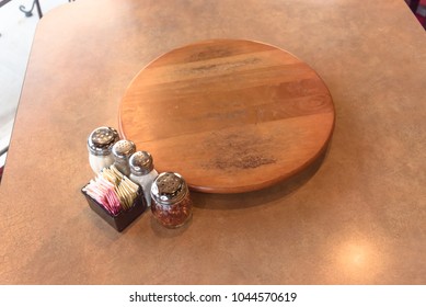 Top View An Empty Wooden Table With Rotating Serving Plate And Shaker Seasonings. Red Pepper Flakes, Parmesan, Black Pepper, Salt