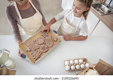 Top view of excited girl taking Christmas cookie from tray. Her grandmother is standing in apron and looking at child with love Stock Photo