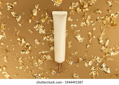 Top View Of Face And Eye Cream Squeeze Cosmetic Tube With Long Nozzle And Bronze Screw Cap And Pieces Of Gold Paper On Gold Background. Cosmetics Spa Branding Mockup, Flatlay.