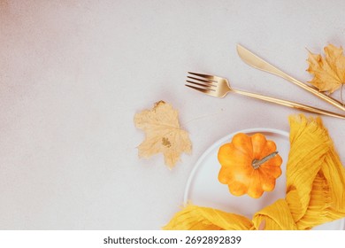 Top view of fall table setting with orange pumpkin, golden cutlery, maple leaves and yellow napkin on light background