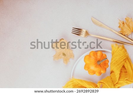 Top view of fall table setting with orange pumpkin, golden cutlery, maple leaves and yellow napkin on light background