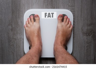 Top View Of The Feet Of A Barefoot Man Standing On Smart Scales, On Whose Balance It Is Written Fat. White Smart Weights With Empty Balance Standing On A Gray Wooden Floor.