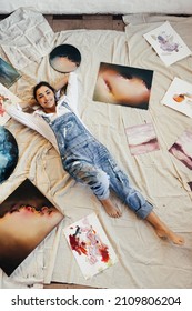 Top view of a female artist lying down in the middle of her paintings. Happy young painter smiling at the camera in her art gallery. Creative young woman displaying a collection of her artwork.