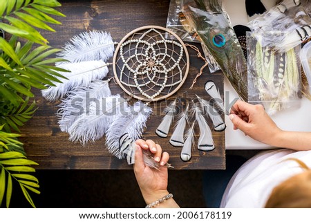 Top view female craftsman hands creating traditional dreamcatcher decor choosing material at workshop. Woman artist making native tribal spiritual amulet use natural feathers, beads and thread