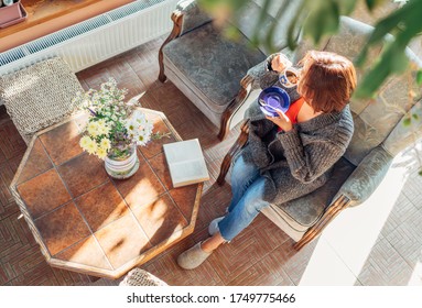 Top View Of A Female Dressed Cozy Home Clothes Sitting In A Comfortable Armchair And Drinking Morning Coffee In House Sunroom Living Room While She Reading A Book.