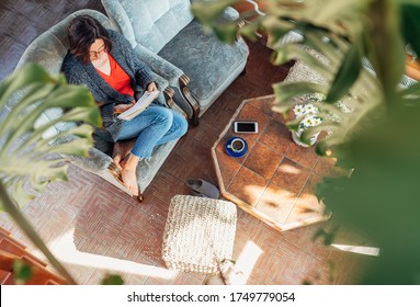 Top View Of A Female Dressed Cozy Home Clothes Sitting In A Comfortable Armchair, Realxing, Reading A Book And Drinking Morning Coffee Or Tea In House Sunroom Living Room. 