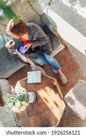 Top View Of A Female Dressed In Cozy Home Clothes Sitting In A Comfortable Armchair And Drinking Morning Coffee In House Sunroom Living Room While She Reading A Book. Stay Home Concept Image.