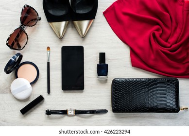 Top View Of Female Fashion Accessories For Woman. Red Clothe, Sunglasses, Face Powder, Make Up Brush, Nail Polish, Watch, Mobile Phone, Lipstick, Gold Pointed Shoes, Black Leather Purse