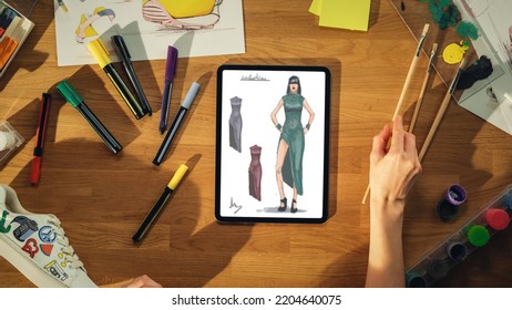 Top View Of Female Fashion Designer Drawing Sketch of Dress on Tablet Computer from Upcoming Collection. Talented Woman Picking Up Paint Brush From the Desk. Clothing Design Process Concept. - Powered by Shutterstock