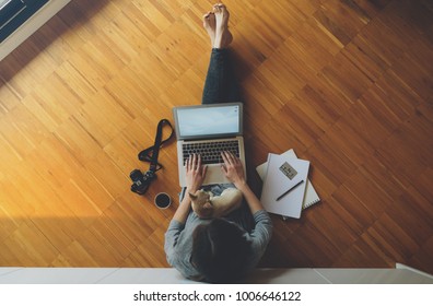 Top View Of A Female Freelance Photographer Working With Laptop Computer, Sitting On A Wooden Floor With Her Little Dog On Her Lap. Girl Looks At Photos From Vacation. Vintage Camera.