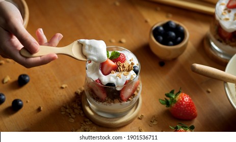 Top View Of Female Hand With Wooden Spoon Eating Granola With Greek Yogurt And Fresh Berries On A Glass