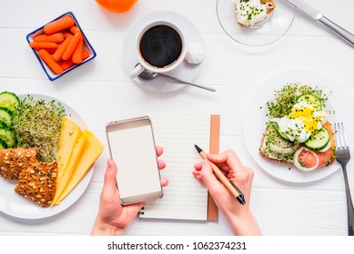Top view Female hands holding smart phone and writing in notebook on served white wooden table with breakfast dishes. Day diet planning and healthy eating concept. Selective focus, copy space - Shutterstock ID 1062374231