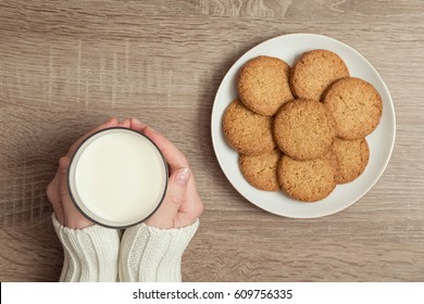 Top View Of Female Hands Holding A Glass Of Milk, With Plate Of Chocolate Chip Cookies Placed Next To It On The Table