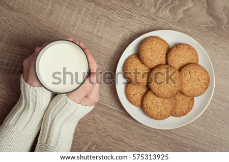 Top view of female hands holding a glass of milk, with plate of chocolate chip cookies placed next to it on the table. Selective focus