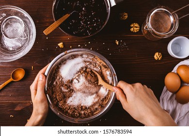 Top view female hands mix cocoa powder, sugar and flour to make dough with melted chocolate and walnuts for delicious homemade brownie cake.