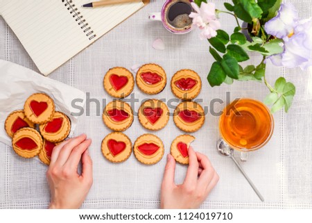 Top view female hands playing tic tac toe game by cookies with hearts and kiss shapes on white table with tea and coffee cups, notebook and fresh flowers. Love concept. Selective focus, copy space.