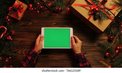 Top View. Female Hands Tapping, Scrolling And Zooming On Tablet Computer Horizontally. The Decorated Wooden Desk With Christmas Lights. Green Screen, Chroma Key.