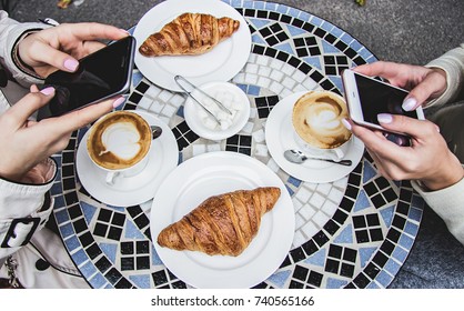 Top view female hands using smart phones while drinking coffee. Two women having coffee brake in French cafe. Girls friends drinking coffee in French styled restaurant. Croissants and coffee on table.