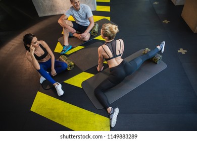 Top View Of Female Instructor Showing Flexibility Exercises To Class. Jolly Man And Woman Are Sitting On Mats And Watching Her. They Are Using Foam Roller For Working On Splits Together