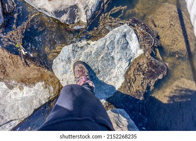 Top View Of A Female Leg In Black Pants And Hiking Boots Walking Across The Stepping Stones Over A River, Calmly Flowing Water With Sunlight Reflecting Shadows