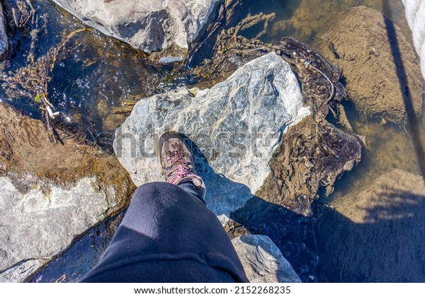 Top view of a female leg in\
black pants and hiking boots walking across the stepping stones\
over a river, calmly flowing water with sunlight reflecting\
shadows