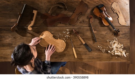 Top view. Female luthier working on the creation of a violin. She sits at a wooden table in her workshop, various tools and instrument are placed on the workbench