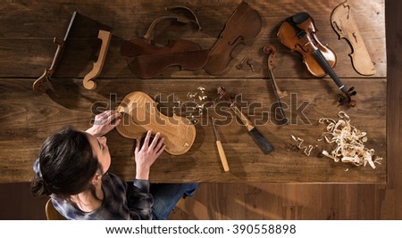 Top view. Female luthier working on the creation of a violin. She sits at a wooden table in her workshop, various tools and instrument are placed on the workbench