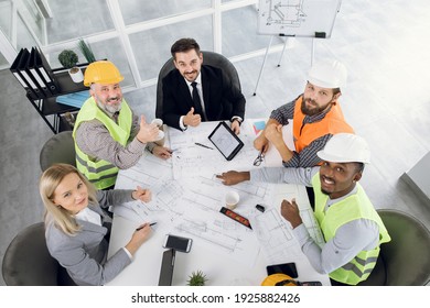 Top View Of Five Diverse People In Suits And Helmets Sitting At Office And Showing Thumbs Up. Qualified Construction Team Planning Arrangement Of Service Lines Of New Building.