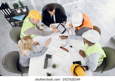 Top View Of Five Diverse People In Suits And Helmets Sitting At Office With Blueprints And Gadgets. Qualified Construction Team Planning Arrangement Of Service Lines Of New Building.