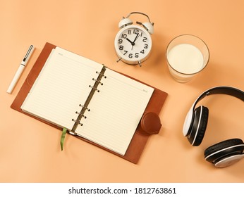 Top View Or Flat Lay Of Alarm Clock ,milk Cup And Headphones With Opened Note Book On Orange Background. Bedtime  Routine Concept.