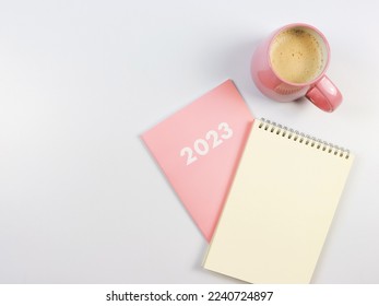 Top view or flat lay of blank paper note book on pink diary 2023 and pink cup of coffee on white background.