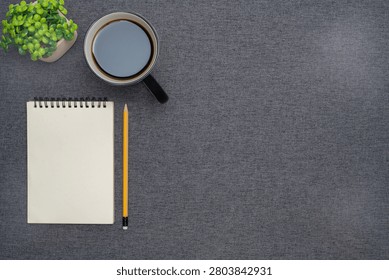 Top view flat lay of a blank spiral notebook, yellow pencil, black coffee cup, and small green plant on a dark grey fabric background with copy space