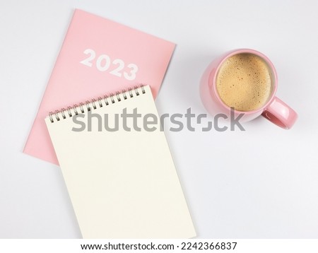 Top view or flat lay of blank paper note book on pink diary 2023 and pink cup of coffee on white background.