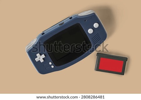 Top view flat lay of a classic dark blue portable handheld video game console with cartridges on beige background. Retro equipment.