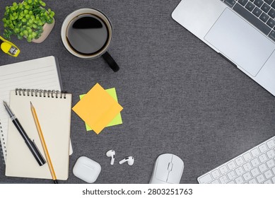 Top view flat lay of a creative home office desk with a laptop, keyboard, mouse, notebook, pens, sticky notes, earbuds, coffee, and plant on a grey fabric background
