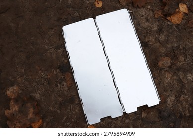 Top view flat lay of folding aluminum camping gas stove windshield screen shield screen protector lying on wet dirt ground with autumn leaves for bushcraft tourist cooking equipment concept