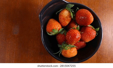 Top view flat lay of fresh, ripe red strawberries in a black bowl on a rustic wooden table. 