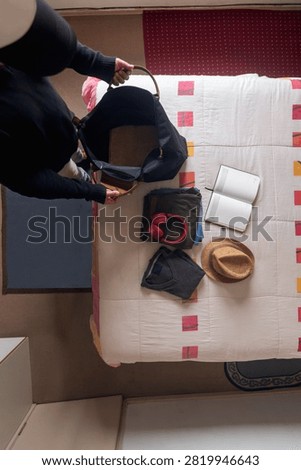 Top view flat lay of a man packing a travel bag on a bed with clothes, headphones, and a hat