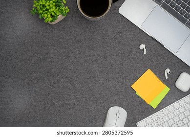 Top view flat lay of a modern office desk workspace with a laptop, keyboard, computer mouse, wireless earbuds, and sticky notes on a dark grey fabric background with copy space